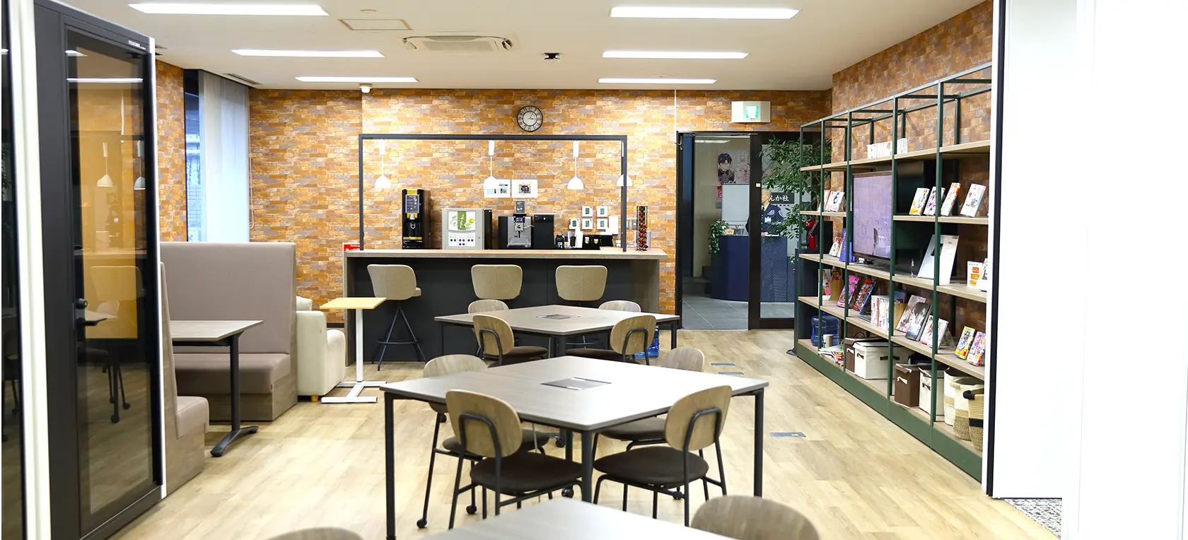 社内風景。テーブルと椅子が並ぶ落ち着いた雰囲気のオープンスペース。壁際には本棚とコーヒーマシンがあり、打ち合わせや休憩に使われている。