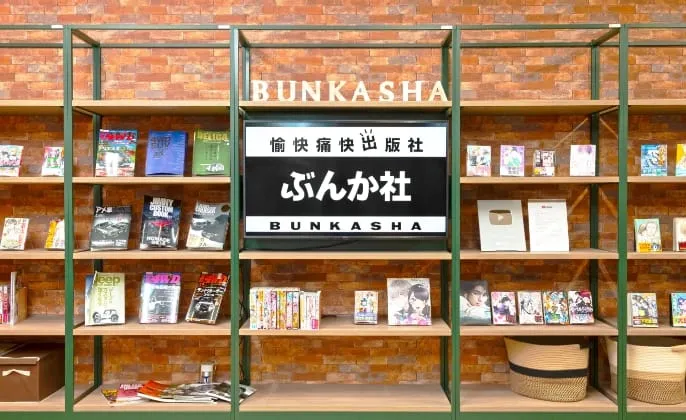 棚に設置されたモニターに『愉快痛快出版社 ぶんか社 BUNKASHA』と表示された、ぶんか社の展示スペース。