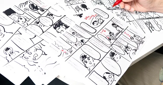漫画のネーム原稿に赤字で修正指示を書き込みながら内容を検討している制作途中の様子。